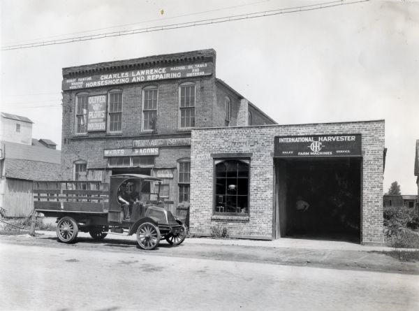 Two men are sitting behind the wheel of a truck with stake body parked in front of an International Harvester dealership storefront. On the right is a one-story building with a large open garage door.