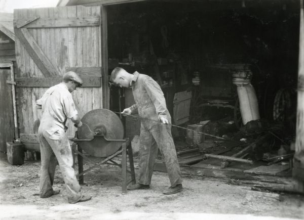 Two men standing near the open doorway of a shed while using a hand-operated knife grinder to sharpen a sickle bar or blade.