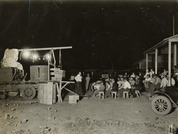 Group gathered on benches in the late evening for a night time lecture, lantern slide show or film showing by International Harvester's Agricultural Extension.