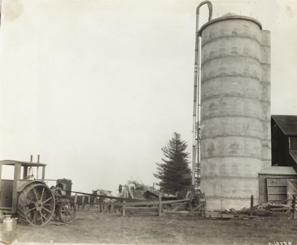 A man is using a silo filler powered by a belt attached to a tractor. Two work horses are tethered on the left, and on the right is a silo and a barn. There is a windmill behind the silo.