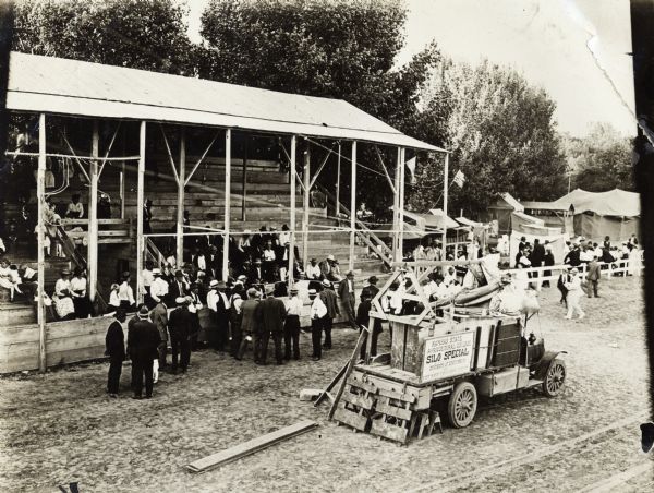 Elevated view of group of people gathered in outdoor pavilion for an exhibition or fair. Truck in the foreground has a sign that reads: "Kansas State Agricultural College Silo Special."