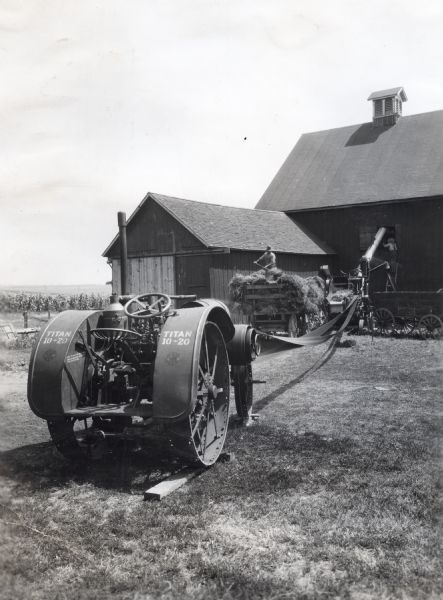 A Titan 10-20 tractor attached to a belt is being used to power an ensilage cutter(?) as men are loading hay from a wagon into the loft of a barn.