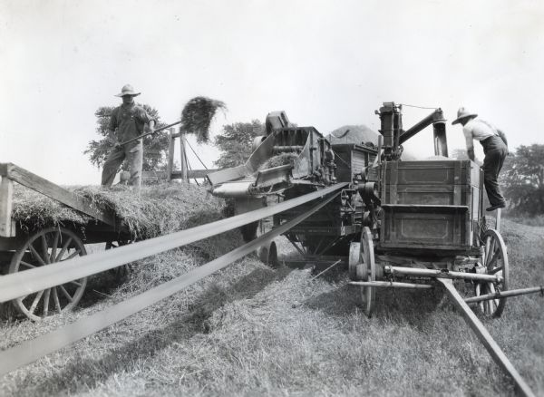 Two men are using a belt-powered hay baler or hay press in a farm field.