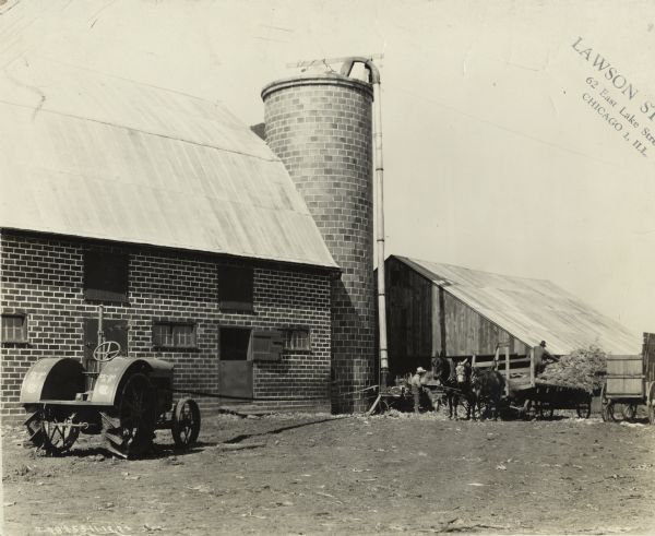 Brick barn and silo. Two men are using a 15-30 tractor to power a silo filler.