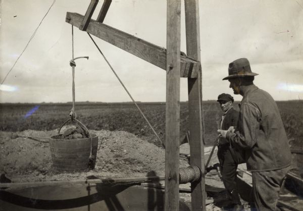 One man is using a hand crank to pull a bucket of earth from the bottom of a pit silo. Another man stands behind him.