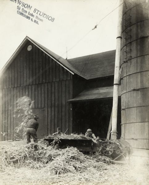 View of a man pitching silage into ensilage cutter and blower near a barn and silo. Two other men are working behind the cutter and blower.