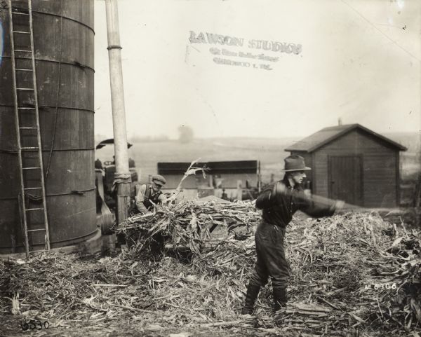 Two men are loading silage into ensilage cutter and blower to fill a silo.