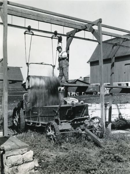 A man is using a pulley on a track system to release manure from a litter carrier into a manure spreader at International Harvester's Hinsdale experimental farm. Barns, silos, and other farm buildings are in the background.