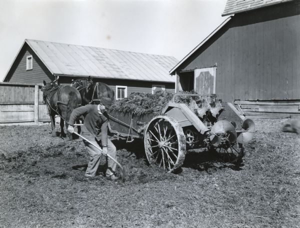 A man is using a shovel to load a McCormick-Deering manure spreader pulled by two horses.