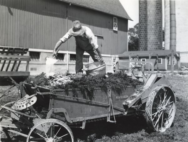 A man standing on the back of a McCormick-Deering manure spreader to pour superphosphate over manure at International Harvester's Hinsdale experimental farm. A barn and silos are in the background.