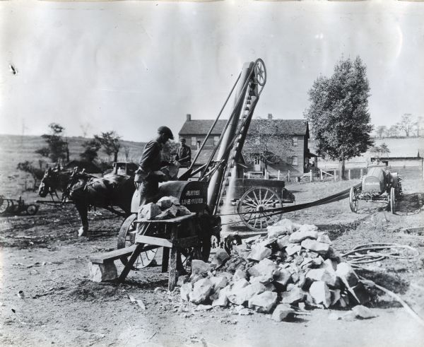 A man is feeding limestone into a "Jeffrey lime pulver" powered by an International tractor. A pile of rocks is in the foreground, and a farmhouse and another man driving a two-horse team are in the background.