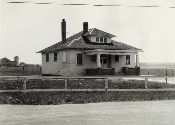 View across road of the front facade of a rural schoolhouse. A small dog is lying on the entrance's front steps, and the sign above reads: "Liberty School District 101." Original caption reads: "Country School Showing Complete Lack of Any Planting - on Dixie Hwy." In the far background is a barn and silo.