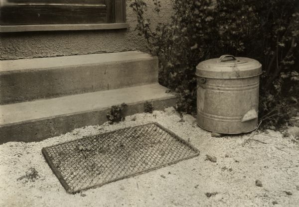 Gravel rear walkway of residence. Small tin container with lid, and a wire mesh mat are set just outside the backdoor steps. Original caption identifies the location as the "Harvester Women's Farm."