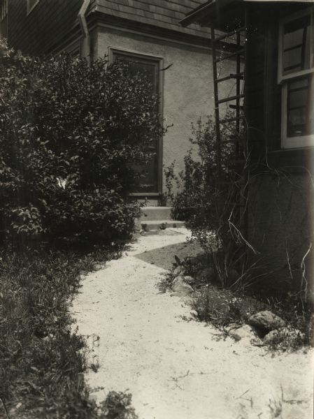 Dirt walkway surrounded by landscaping to door of residence. The original caption identifies the location as the "Harvester Women's Farm."