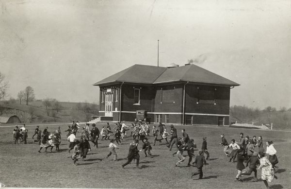 Elevated view of school children running and playing on open school grounds. There is a schoolhouse in the background.
