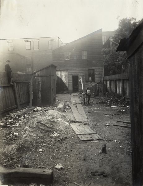 Three young children standing in a backyard in very poor condition, including the lack of a lawn, exposed trash piles, and other litter.