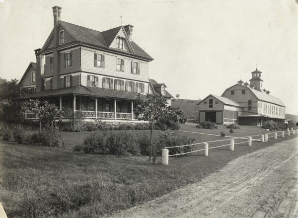 View from a rural road to a farmhouse demonstrating its landscaping and what the original caption describes as a "novel fence." The large barn in the background has a weather vane on top of a cupola, and six ornate roof vents.