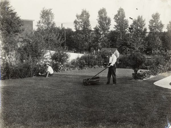 A man and woman (possibly husband and wife) maintain a residential backyard. The man, wearing a bow tie, is mowing the lawn with a reel mower, while the woman, wearing a hat, is sitting on the grass holding small pruning shears for shrubs near a fence.