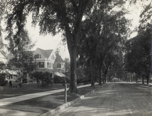 View down a wide, tree-lined neighborhood street. Well-maintained lawns and houses can be seen on the left. A man with a reel mower is working in a yard, and children are playing in the street in the far background.