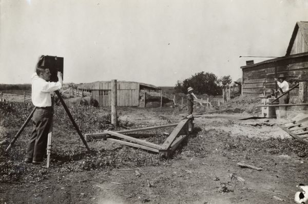 A man uses a camera mounted on a tripod to film another man and two boys near a farm building. The man and boys appear to be constructing a silo. Two horses stand in the background.