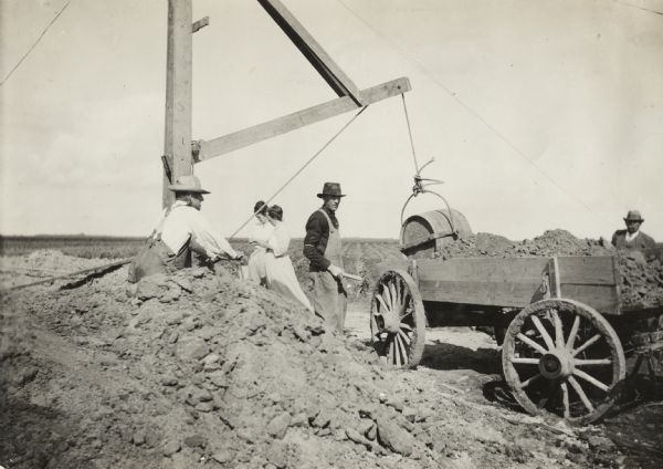Men loading dirt into a wagon while digging for a new pit silo using a derrick. Two women are standing in the background.