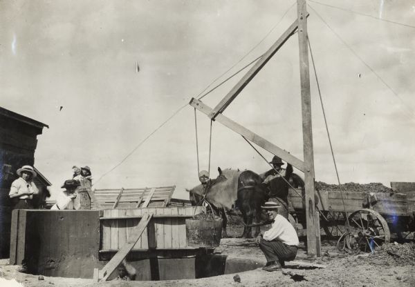 Men and women at the construction site for new pit silo. One man is squatting at the edge of the pit near the derrick, while two women are standing behind one of the completed sections of concrete curbs. One man is working inside the pit.