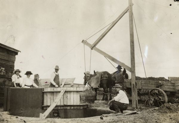 Men and women at the construction site for a new pit silo. One man is squatting at the edge of the pit near a derrick, while two women are standing behind one of the completed sections of concrete curbs. Another man wearing a hat is standing inside the pit. A wooden form still covers one section of concrete.
