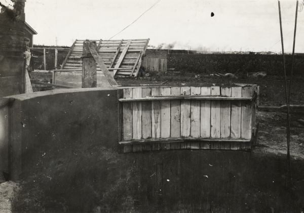 View of the construction of the concrete curbs of pit silo, including a wooden form. On the left is a young girl looking out from behind a fence. In the far background beyond a field a train is going by a town in the far background.
