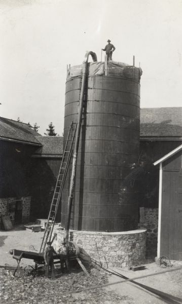 Elevated view of two men in a barnyard with a silo filler. Another man is standing on top of the silo.