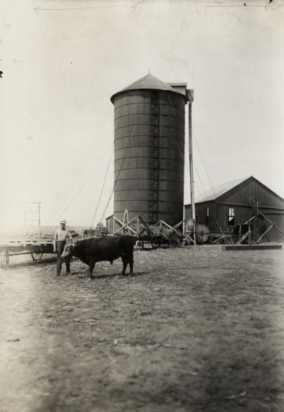 A farmer wearing hat is standing in the middle distance beside a bull. Behind him is a large silo.