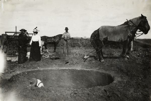 Two men and women at the construction site for new silo. One man holds the reins for nearby team of work horses while the other man stands inside the pit for the pit. Two well-dressed women stand at the edge of the silo.