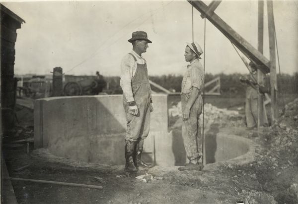 Two workers talking while standing at the site for a new silo. The pit and foundation for the silo is in the background. A third man is standing near a derrick at the edge of the pit.