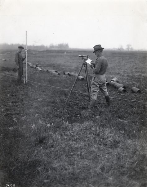 A.L. Webster takes notes in a notebook while using tools to do surveying work in a field. Another man stands holding a rod in the background near what appears to be cut segments of pipe.
