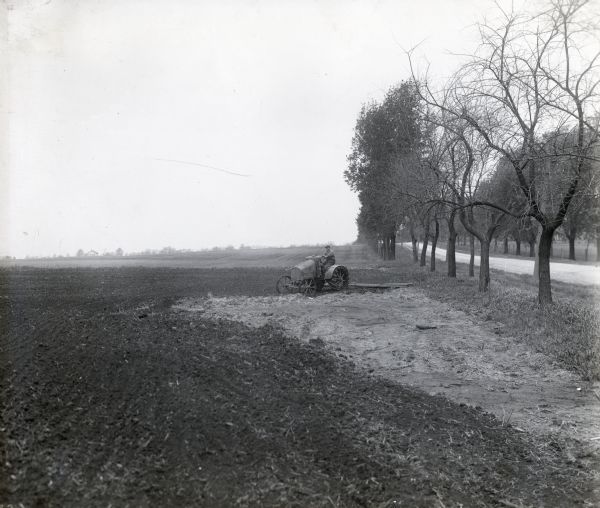 A man is operating an International 8-16 tractor around a wet area in a field at International Harvester's Hinsdale experimental farm. On the right is a tree-lined road.
