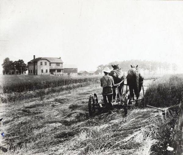 Rear view of a man leading two horses pulling a mower across a field. In the background is a farmhouse, a barn, and several other farm buildings.