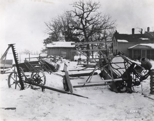 Several farm implements, including a grain binder and mower, sitting in a barnyard covered with snow. A shed, farmhouse, and woodpile are in the background. The photograph was likely taken to demonstrate the hazards of improperly storing equipment.