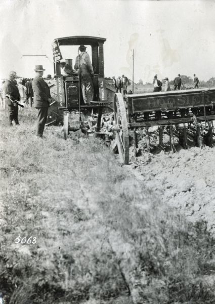Men are watching the demonstration of an Avery Company tractor pulling a Hoosier seed drill (?) across a field at Newsorn Farm during the annual meeting of the Agricultural Extension Commission of the National Implement and Vehicle Association.