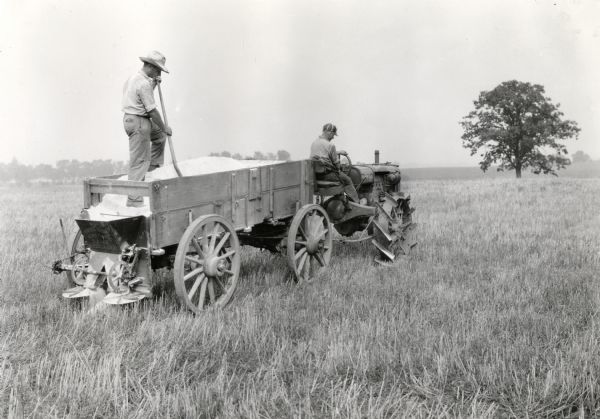 A man standing in an endgate lime spreader is using a shovel to spread lime onto a field at International Harvester's Hinsdale experimental farm. The spreader is being pulled by another man sitting on a Farmall Regular tractor.