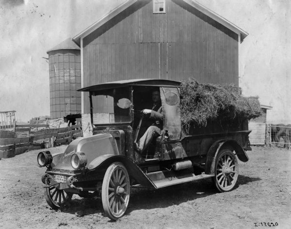 Three-quarter view from front left of a man sitting behind the wheel of an International truck parked in front of farm buildings. The truck is loaded with what appears to be hay. The side of the truck is labeled: "Matt Murphy."