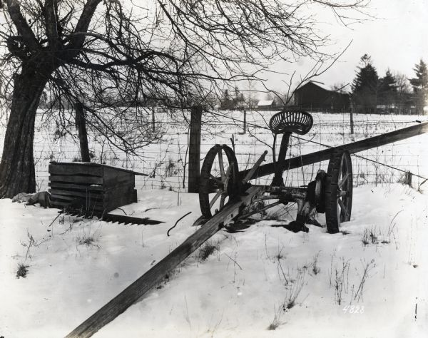 A sickle bar mower is sitting in snow beside a wire fence, illustrating the hazards of improper equipment housing. A barn and other farm buildings are in the background.