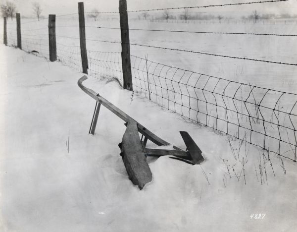 A walking plow resting on its side is on snow-covered ground beside a wire fence. The photograph was probably taken to illustrate the hazards of improper equipment storage.