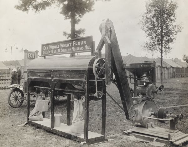 A Meadows stone burr grain mill powered by an International Harvester engine grinding wheat into flour at what appears to be an outdoor fair or exhibition. The sign above the mill reads, "Eat Whole Wheat Flour; Ground with an I.H.C. Engine and Meadows Mill."