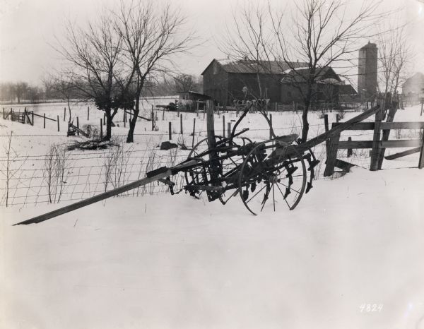 A cultivator sitting in the snow near a fence, illustrating the hazards of improper equipment housing. A herd of cows is in the background near a barn and silo.