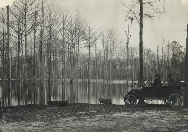 Three men in an automobile near what may be a flooded area, or possibly an overflowed pond or river. Barren trees stand in the pool's water.