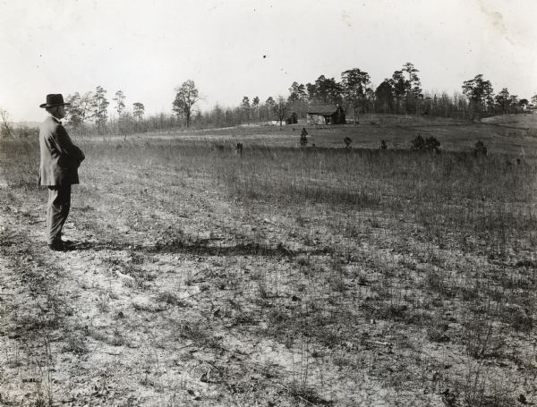 Lindsey Reese, dressed in a hat and suit jacket, standing in an abandoned field. Several farm buildings are in the far background.
