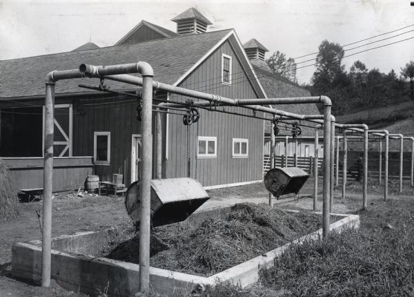 A concrete manure pit and litter carriers operated on pulleys stand in front of multiple farm buildings.