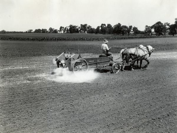 A man is driving a team of two horses across a field that are pulling a McCormick-Deering manure spreader with a lime spreader attachment at International Harvester's Hinsdale experimental farm.