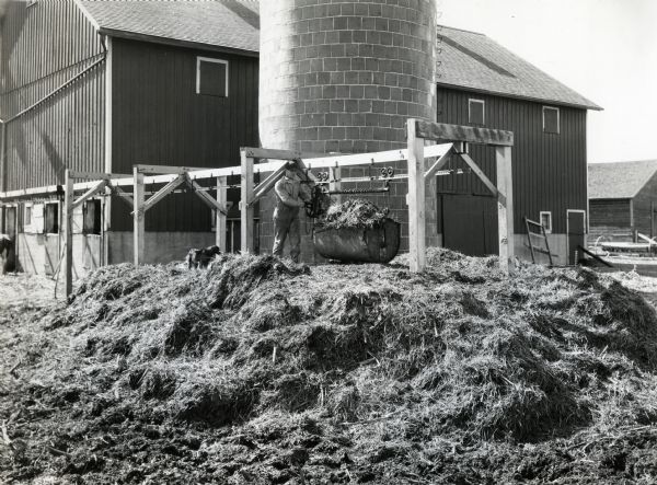 J. Williams, wearing a baseball cap and coveralls, loading a litter carrier next to a manure pile in a barnyard. A dog is standing near him, and a silo and barn are behind them.