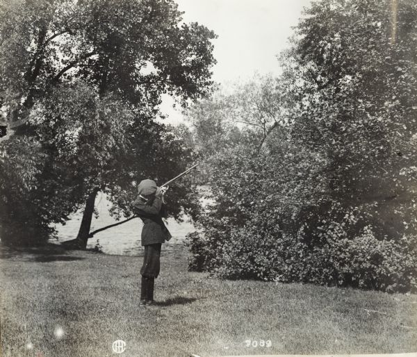 Young boy standing on lawn aiming shotgun into trees. In the background is a lake or river.