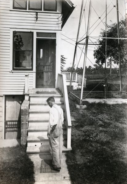 A man wearing overalls standing on a walkway leading to the back entrance of Professor Holden's farm. The base of what appears to be a windmill is to the right of the house.